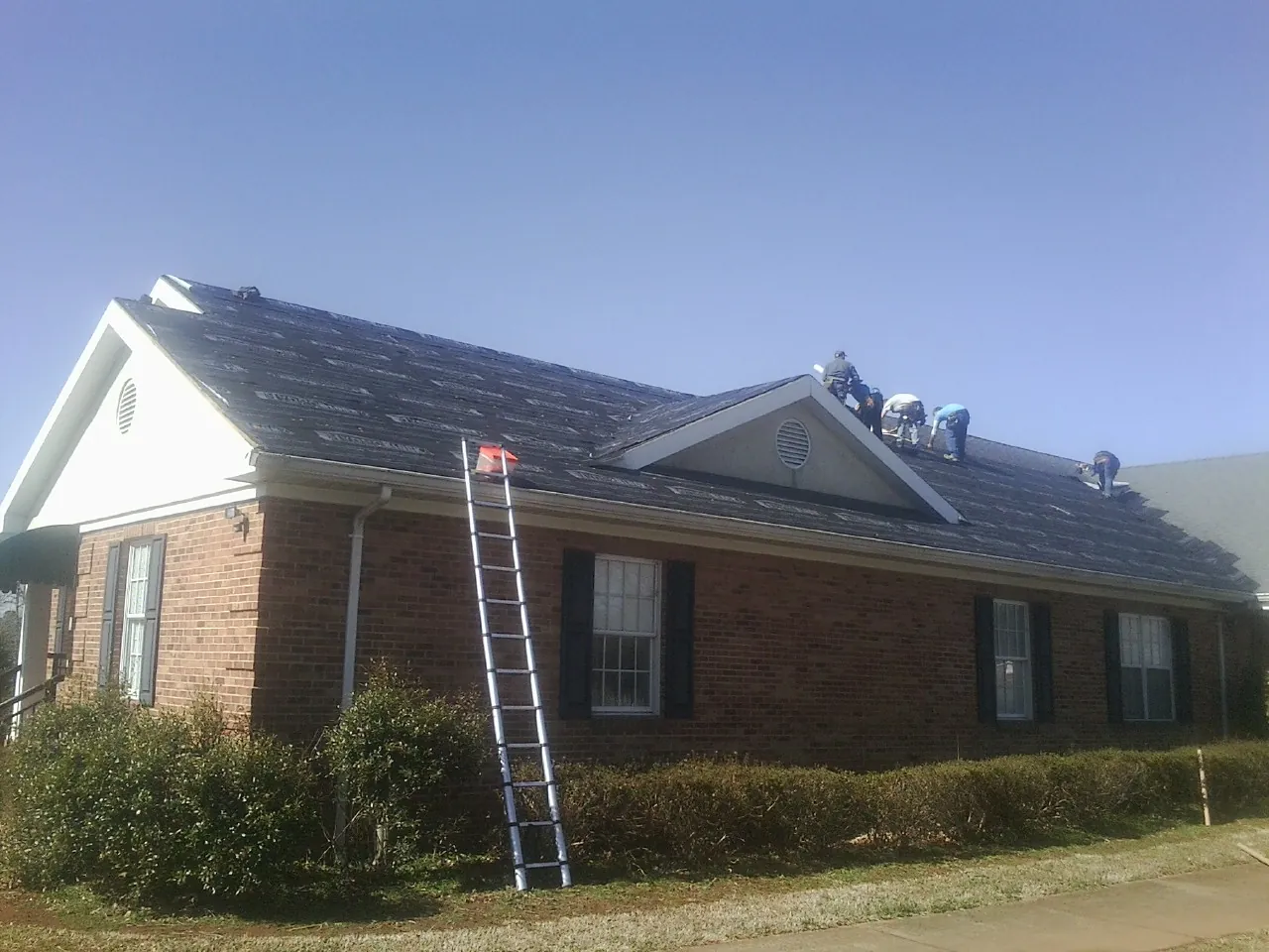 Crew tearing shingles off a brick ranch roof with a ladder set — Emergency Roof Repair work by our crew in Flat Rock