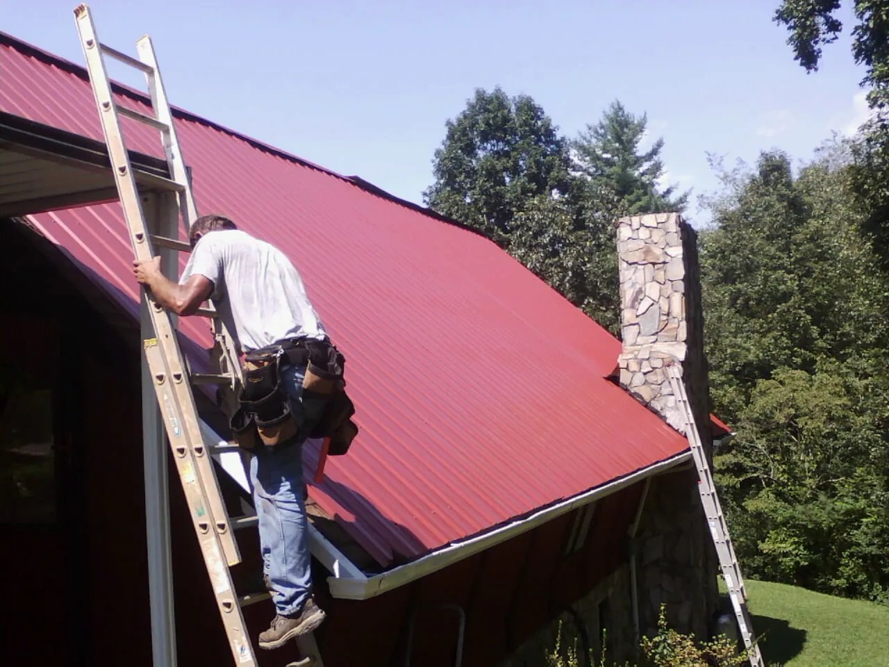 Roofer on a ladder beside a red metal roof and stone chimney — Soffit & Fascia Repair by Roofing Brothers in Flat Rock